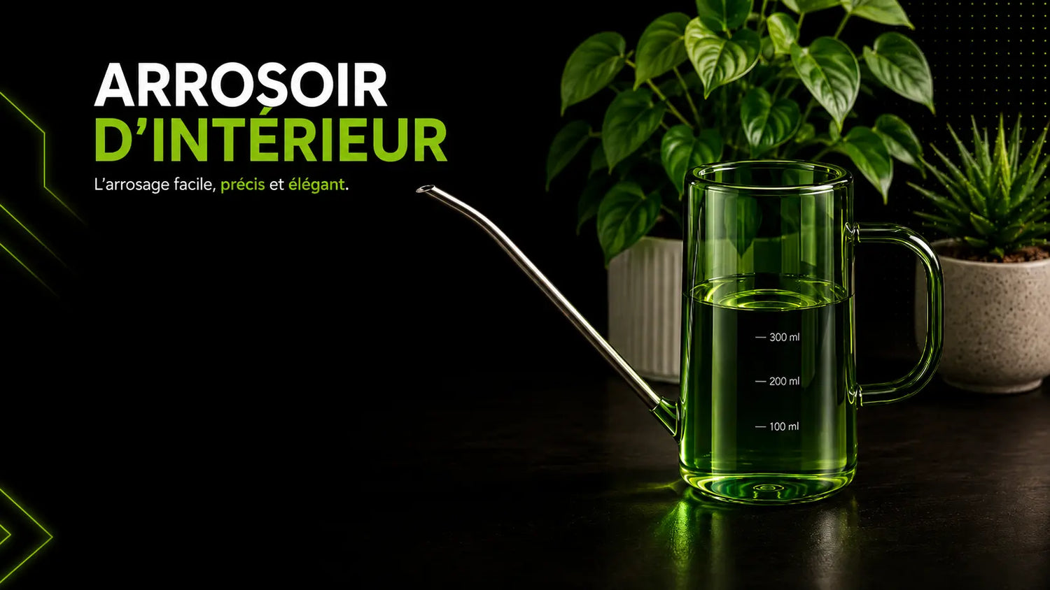 Arrosoir d'intérieur en verre transparent avec un long bec, posé sur une surface sombre près de plantes vertes