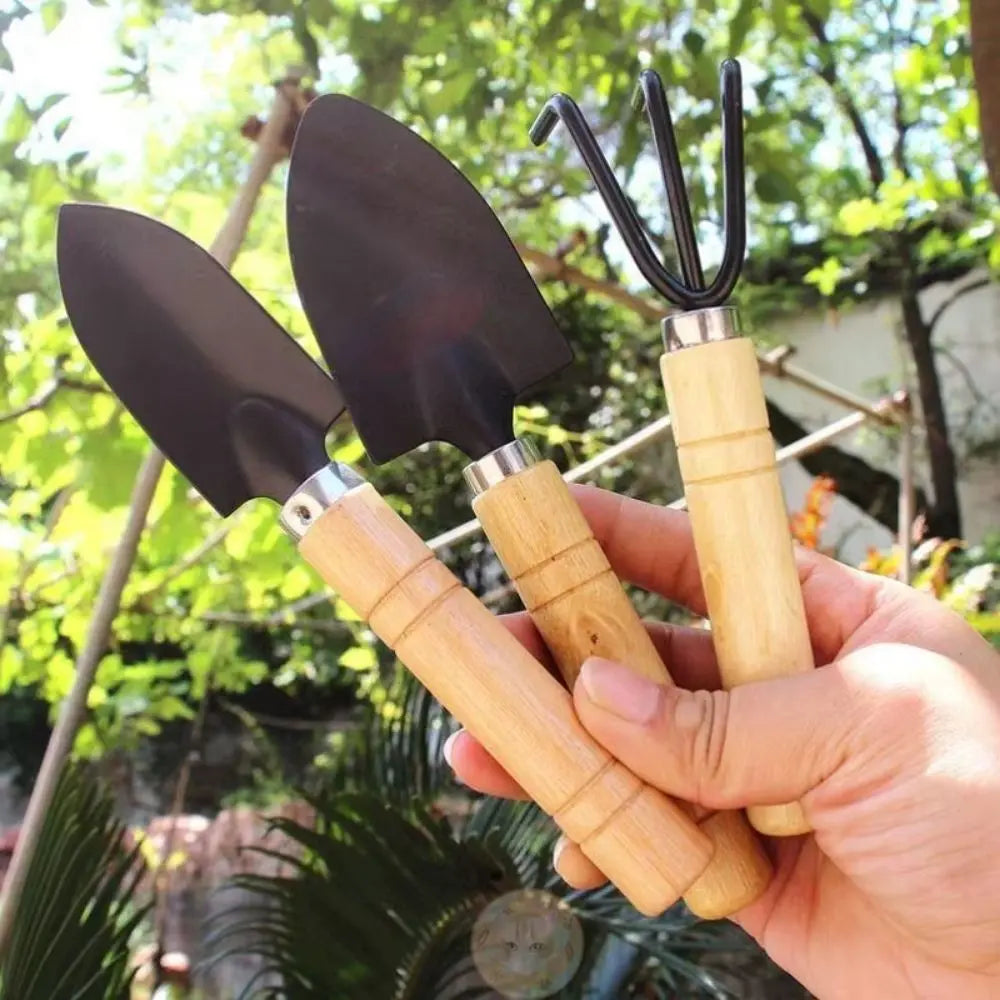 Trois outils de jardinage avec manches en bois et têtes en métal, sur fond de verdure