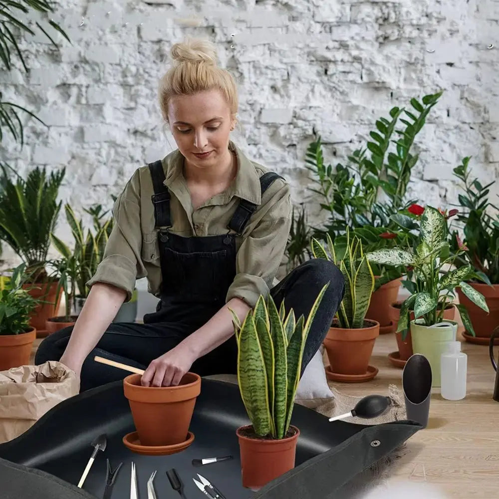 Femme en salopette noire rempotant une plante sur un tapis de rempotage en PVC, entourée de pots et de verdure
